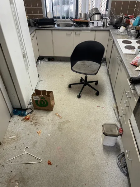 A kitchen floor covered in spilled food, a cardboard box, a coat hanger and a chair, with the benches stacked with pots and dishes.