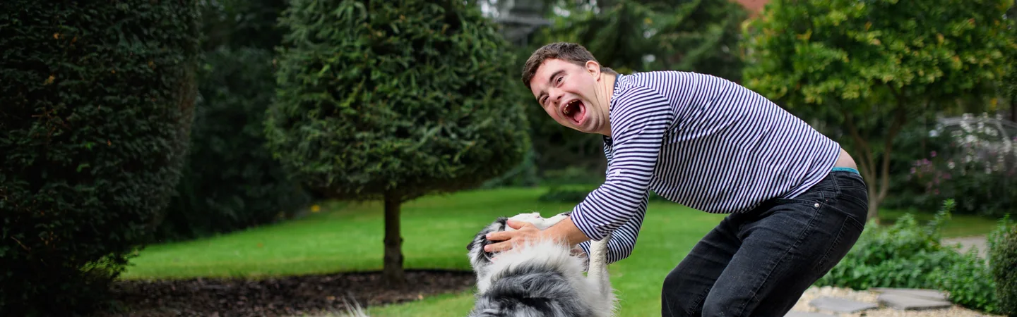 A young man laughing while playing with his fluffy dog in a tidy garden.