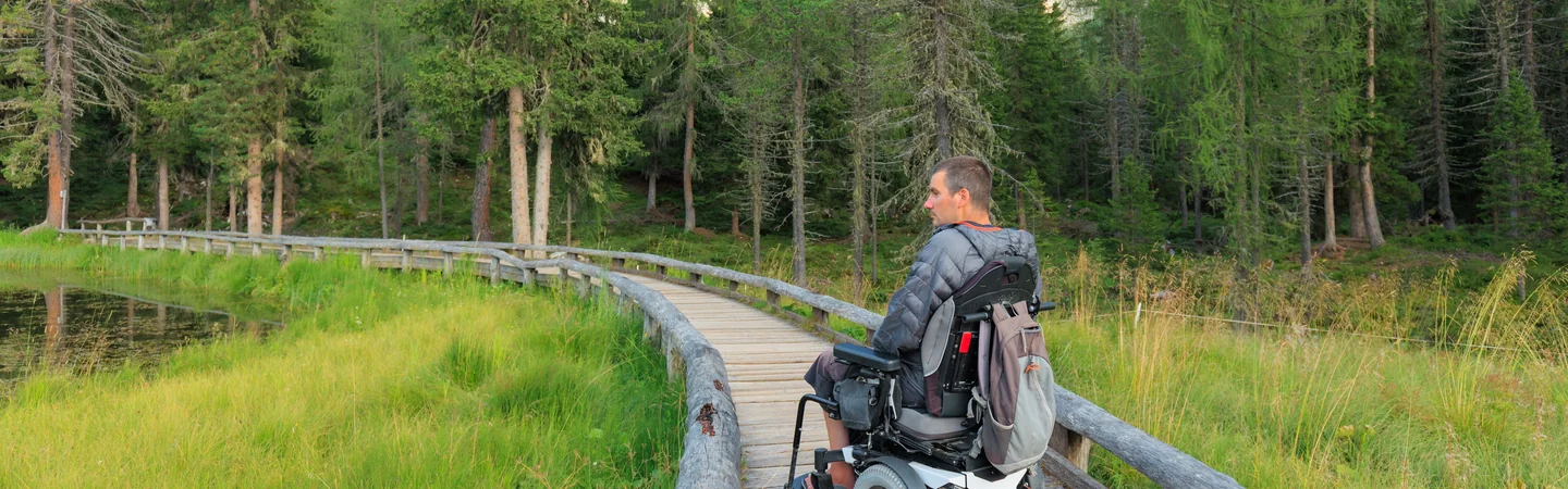 A man in a powered wheelchair on a timber boardwalk through wetland and forest.