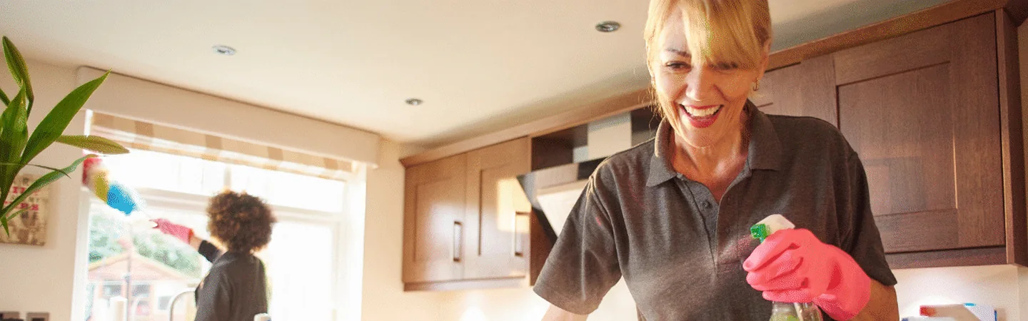 A smiling cleaner holding a spray bottle in a bright kitchen, a colleague dusting the window behind her.