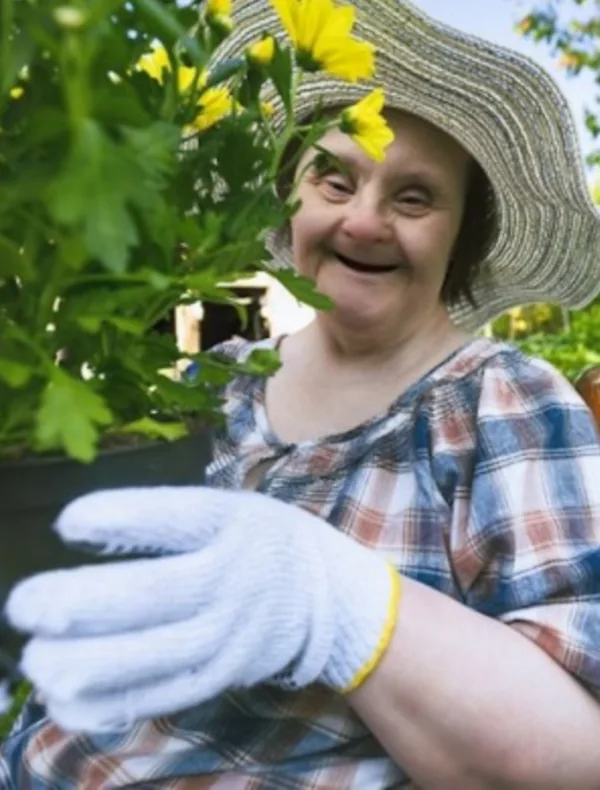 A smiling woman in a sun hat and gardening gloves tending a pot of yellow
                  flowers.