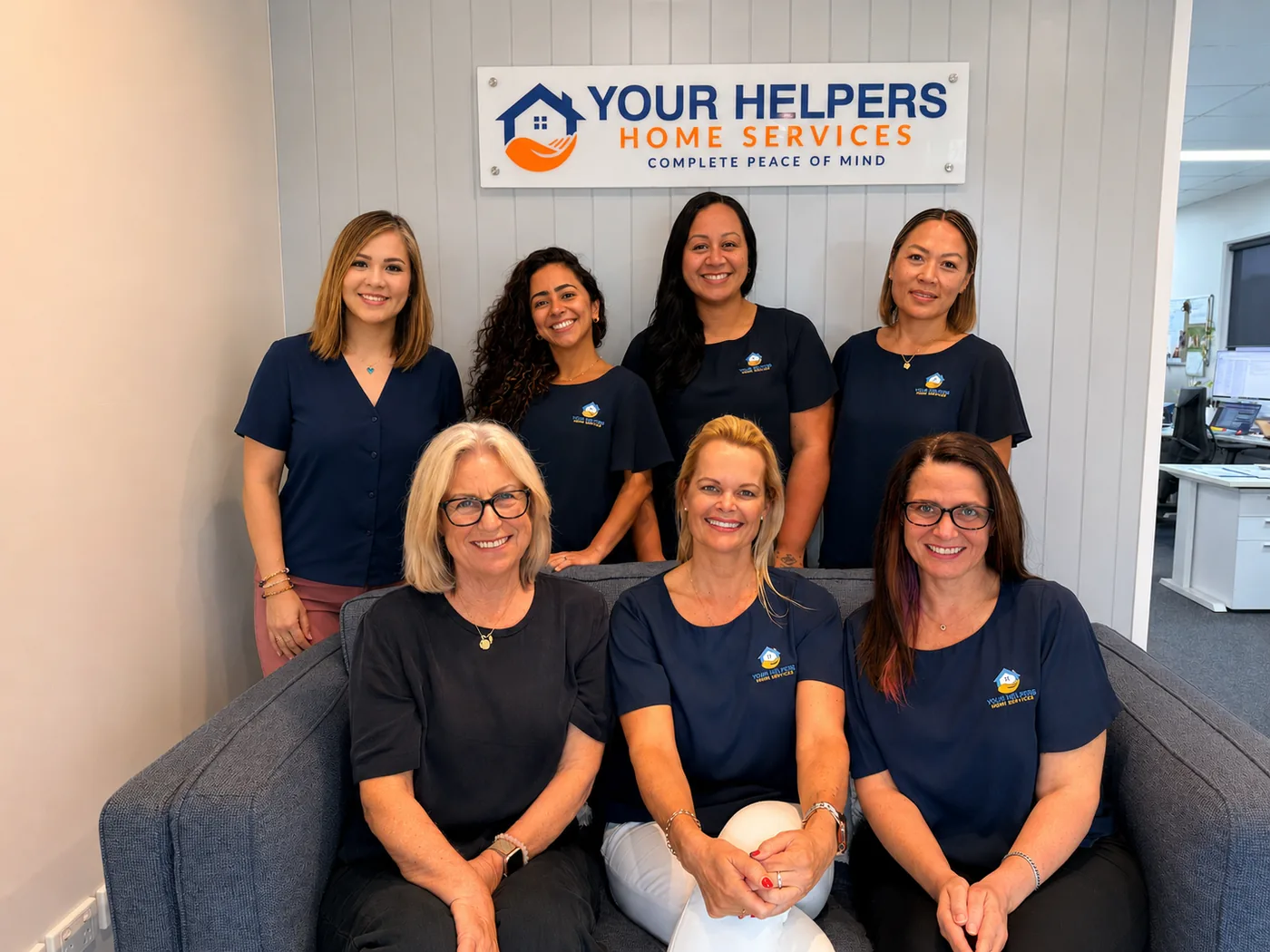 The Your Helpers office team of seven in navy branded shirts, together beneath
                the Complete Peace of Mind sign at the Oxenford office.